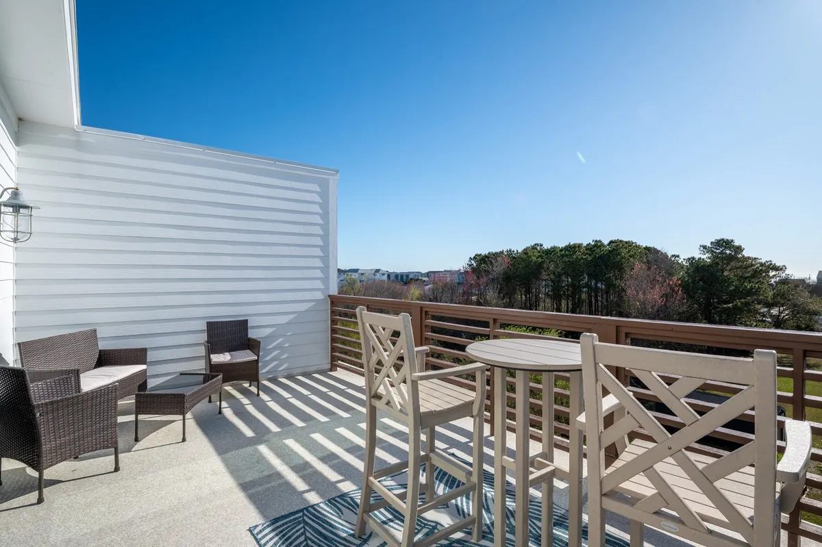 A bistro table with stylish outdoor chairs sits on a rooftop deck, surrounded by railings that overlook lush island landscape and the distant ocean. The scene is set for morning coffee, reading, or sunset viewing, and the relaxed arrangement invites guests to linger and soak in the views.