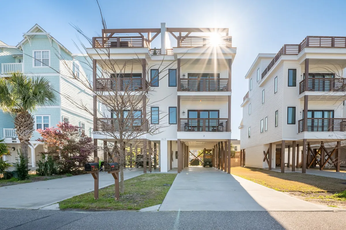 The image showcases a modern single-family house with striking architectural lines, multiple sunlit balconies, and inviting outdoor living spaces. Sunshine glints off the fresh white facade, hinting at the coastal setting. The balconies are set up for relaxation, offering spots for yoga at sunrise, gatherings for evening barbecues, and serene stargazing, all just a short walk from Carolina Beach and the lake.
