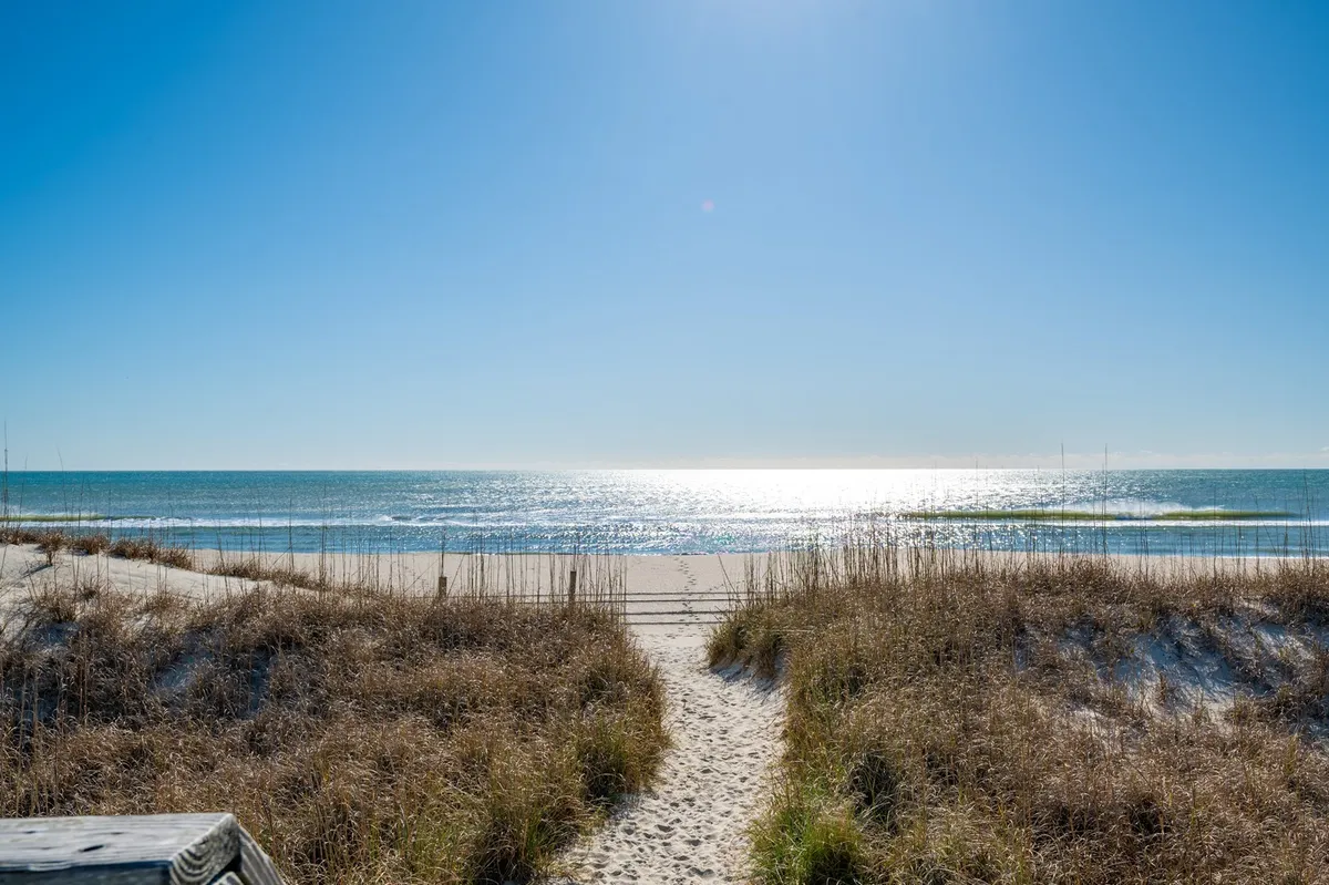 A serene sandy pathway leads through dune grass directly to the expansive shoreline, with gentle waves rolling onto the beach. The sky is clear, and the open space invites play, relaxation, and exploration.