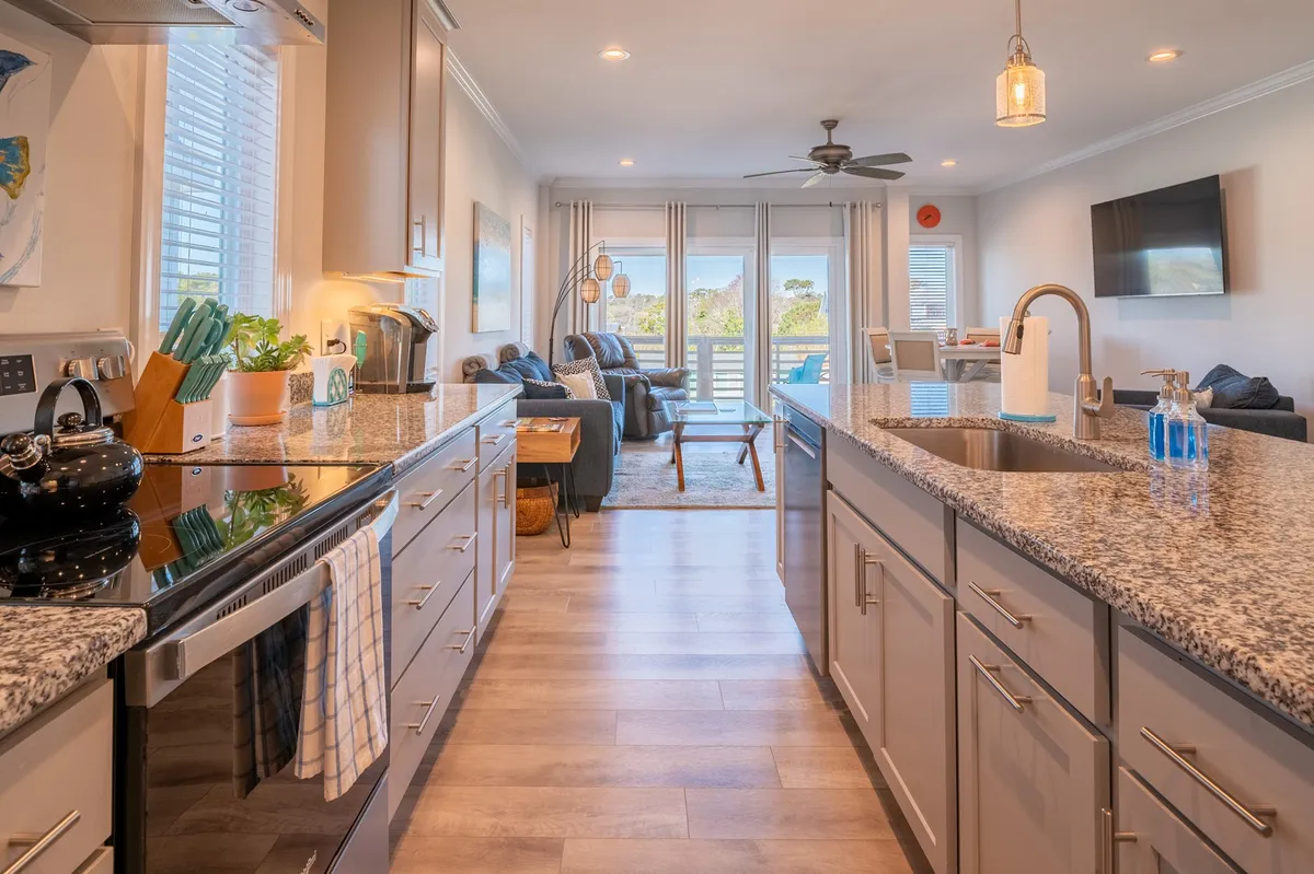 A welcoming, sunlit open-concept living area featuring granite countertops, black appliances, an airy kitchen island, and a comfortable seating area. Large windows let in abundant natural light, and the layout flows smoothly from kitchen to living room, with balcony access visible in the background.