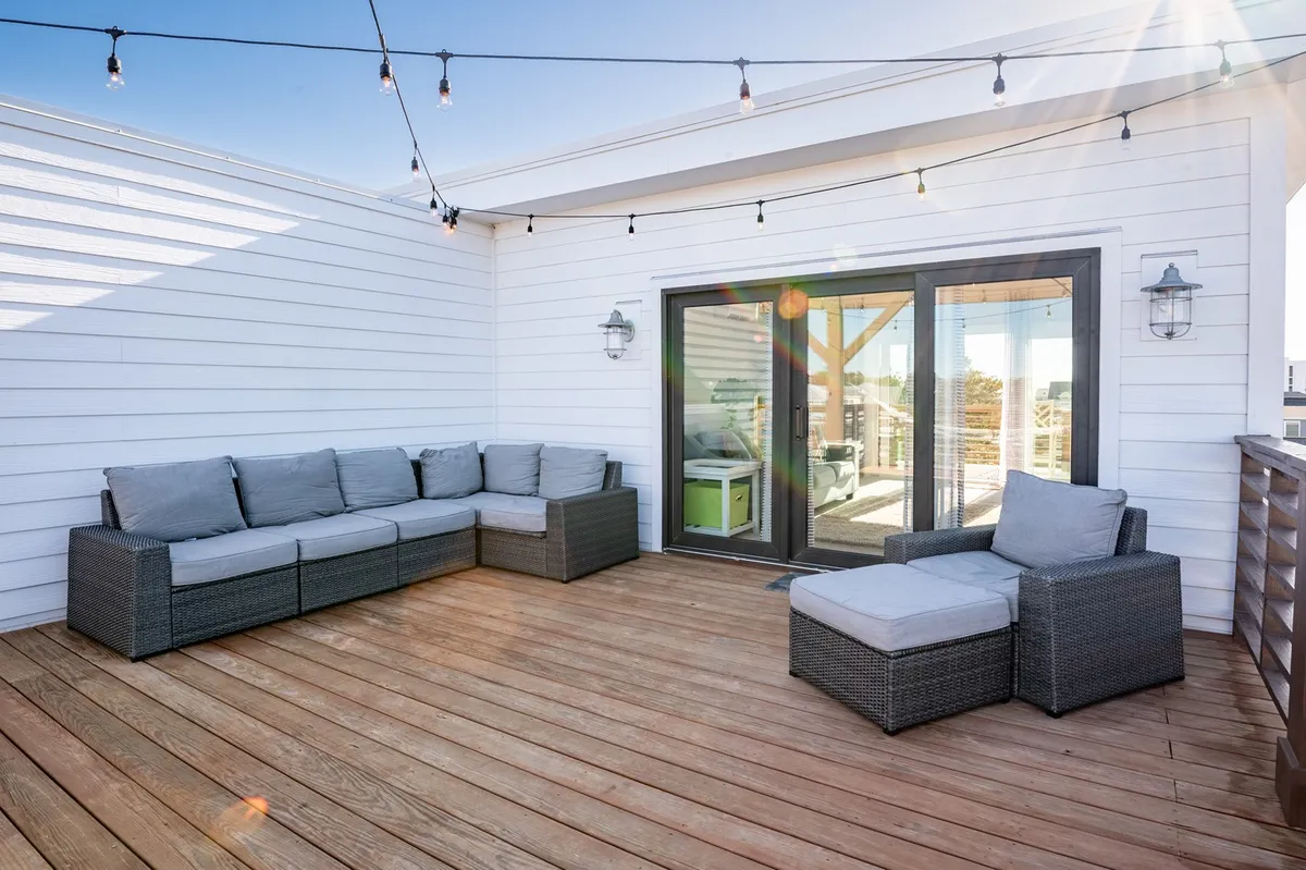 A rooftop deck scene with a plush sectional sofa and outdoor chair, illuminated by string lights that create a warm, inviting glow. Glass doors connect the deck to the interior, and the setup is clearly designed for both relaxation and socializing while enjoying expansive views—ideal for sunset cocktails or evening chats.