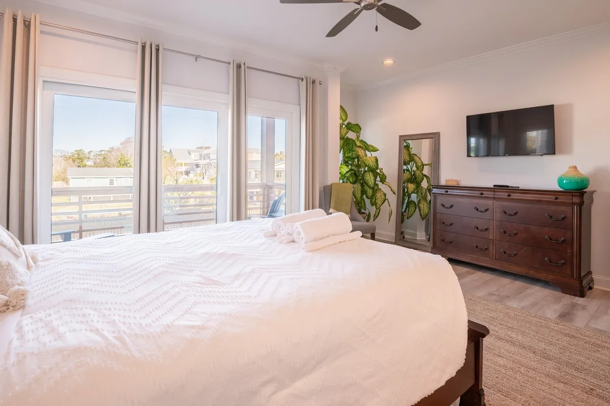 A sunlit bedroom featuring a bed made up with crisp white linens, large windows letting in natural light, a decorative plant, a wooden dresser, and a wall-mounted smart TV. The atmosphere is tranquil and welcoming, perfect for rest and relaxation.