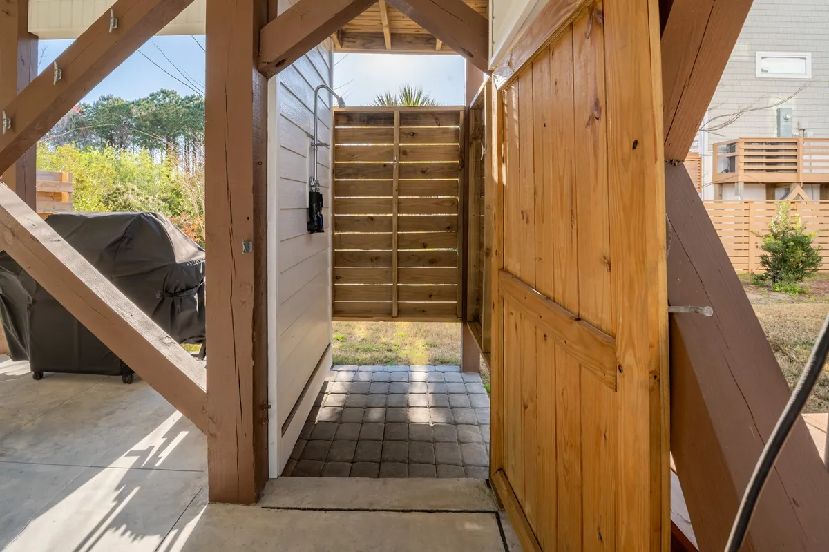The image features an outdoor shower set on a charming patio area, with a wooden door and greenery in the background. It's a dedicated space for rinsing off after a beach day and visually evokes the transition from sandy adventures to comfortable home life.