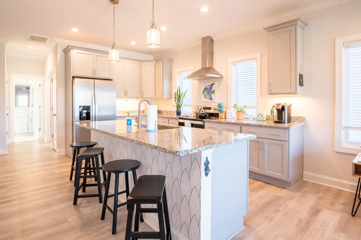 A warm, inviting kitchen scene highlighting the granite kitchen island, stainless steel appliances, bar stools for casual seating, and window views. The layout encourages social cooking and conversation, fitting the modern coastal vibe.