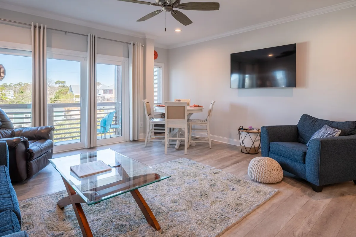 A cozy seating area with a glass coffee table, comfortable chairs, a dining table, and large windows. The room is filled with natural light, suggesting a welcoming environment for both social gatherings and quiet relaxation. The windows suggest a connection to the surrounding area and the potential for views of local festivities or serene landscapes.