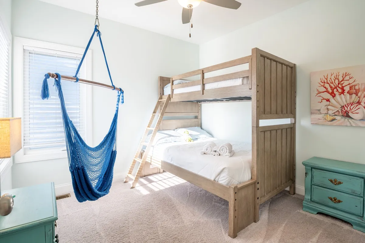 A playful family guest room with a twin-over-full bunk bed, a cozy hammock, decorative wall art, nightstand with a lamp, and plenty of sunlight filtering in. The setup feels inviting for kids and families alike, encouraging both play and relaxation.