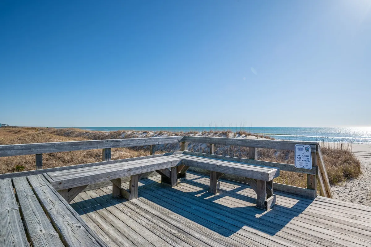 This exterior image features wooden benches overlooking the ocean, with sand dunes and a sign in the foreground. The scene invites viewers to relax, gather, and take in the natural beauty of the coast, with the open sky and tranquil water emphasizing a peaceful, outdoor experience.