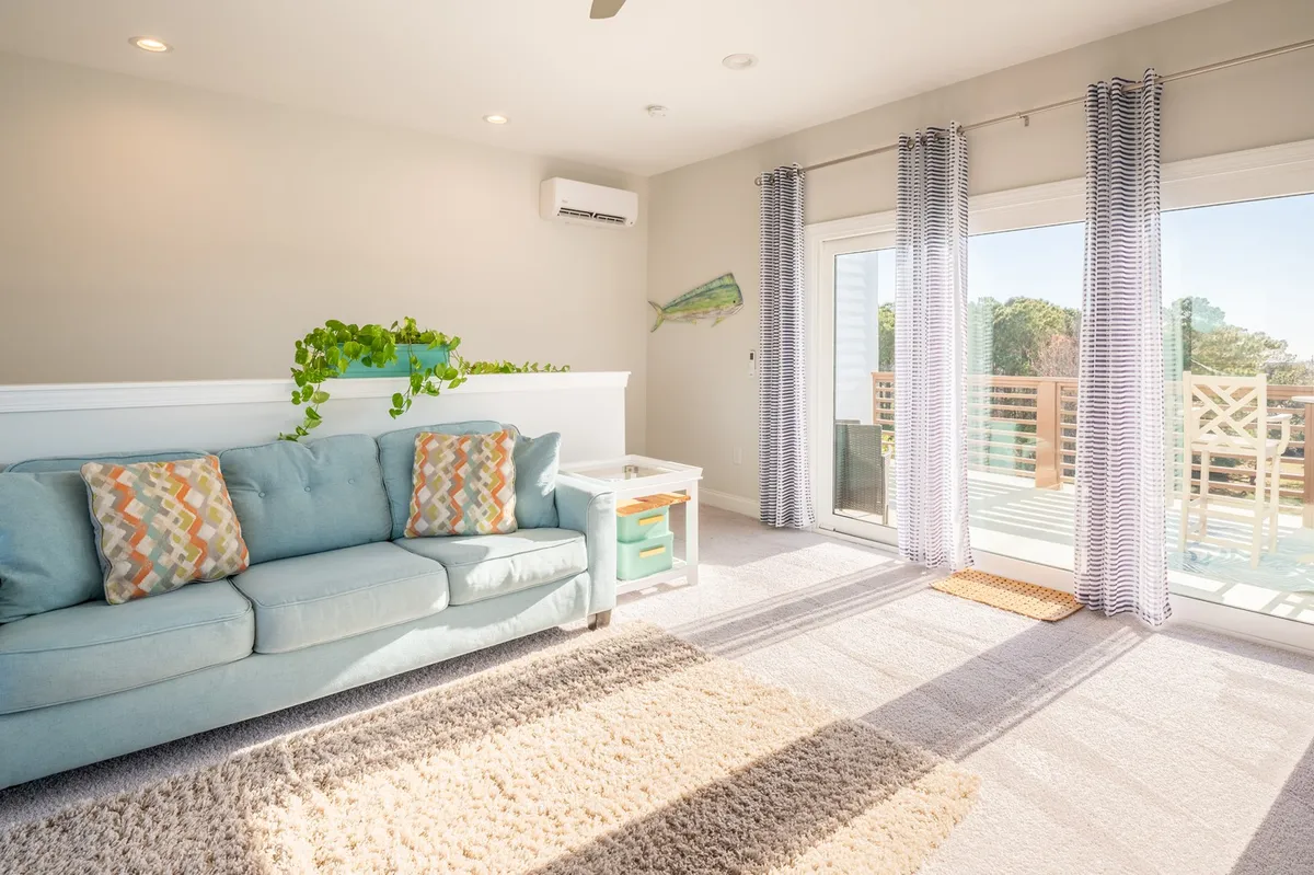 A light-filled living area with a light blue sofa adorned with colorful throw pillows sits next to large windows that look out onto an outdoor deck. Sunlight pours in, and the space feels cheerful and connected to nature. The view of the outdoor deck hints at opportunities for enjoying fresh air, whether it's lively summer gatherings or peaceful off-season relaxation.