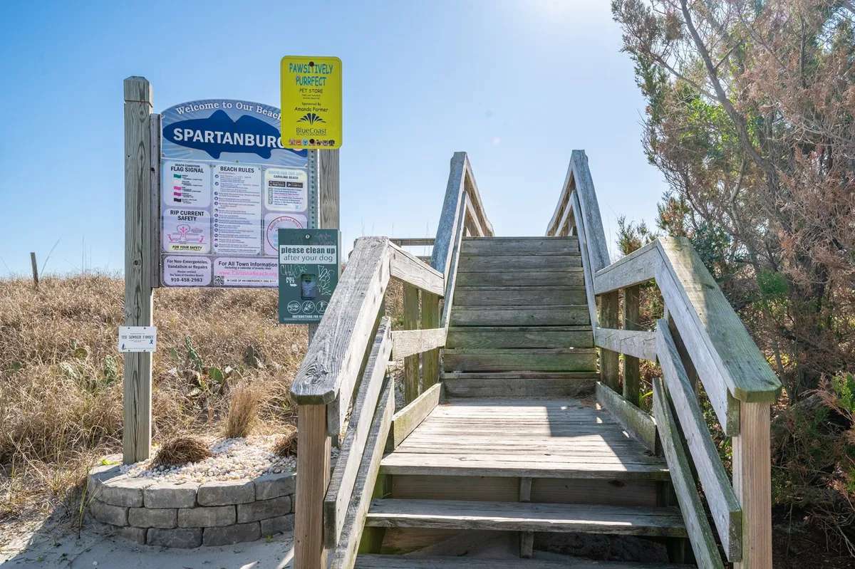 Wooden steps descend through lush greenery, past an informative sign, leading towards the beach. The surrounding vegetation and inviting pathway hint at adventures ahead.