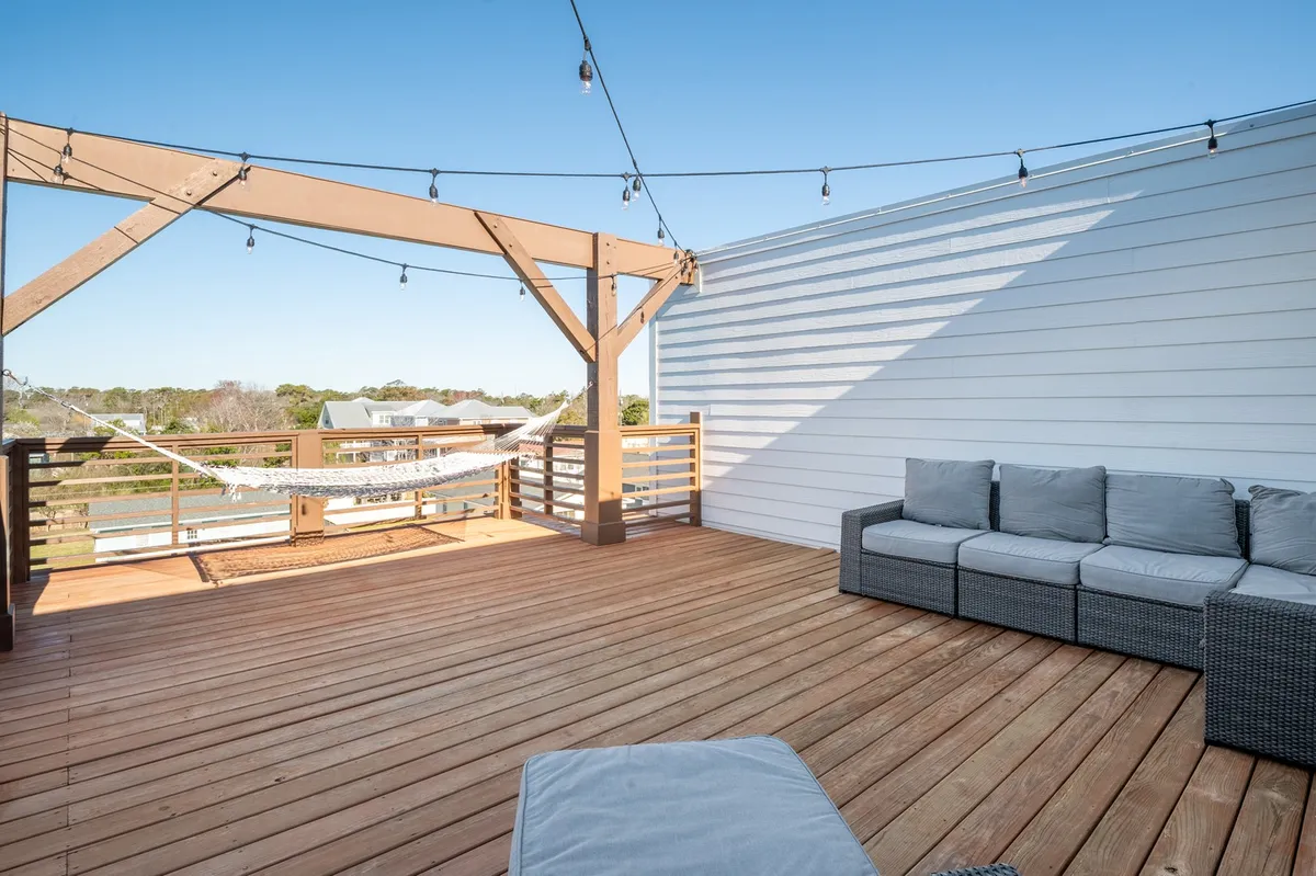 A tranquil rooftop deck scene featuring a hammock strung between posts, an inviting outdoor seating area, and soft string lights. The deck is surrounded by railings, giving a sense of elevation and openness, with glimpses of the ocean and sky in the background. The cozy, relaxed arrangement hints at both daytime lounging and magical evenings under the stars.