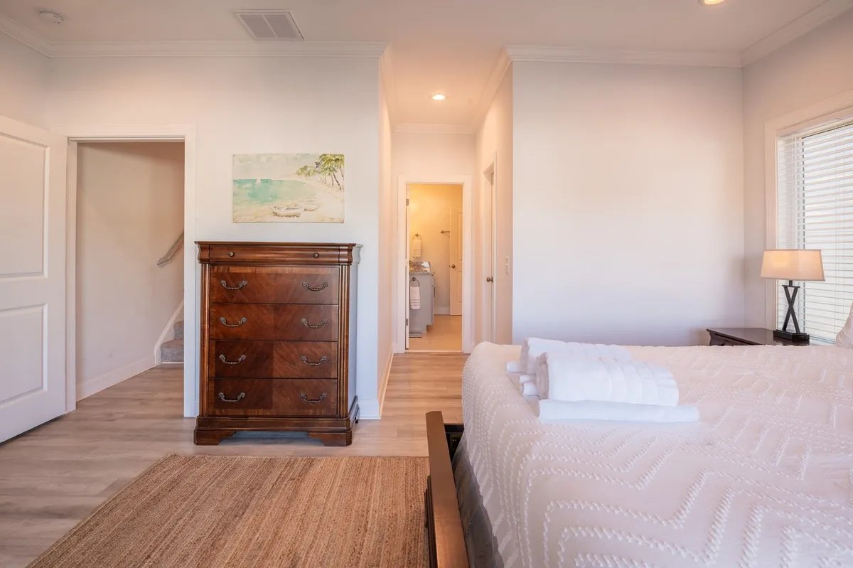 A cozy bedroom with a neatly arranged bed topped with fresh linens, a wooden dresser, bedside lamp, and tasteful wall art. Sunlight streams in, creating a warm and welcoming environment ideal for unwinding after a day of activity.