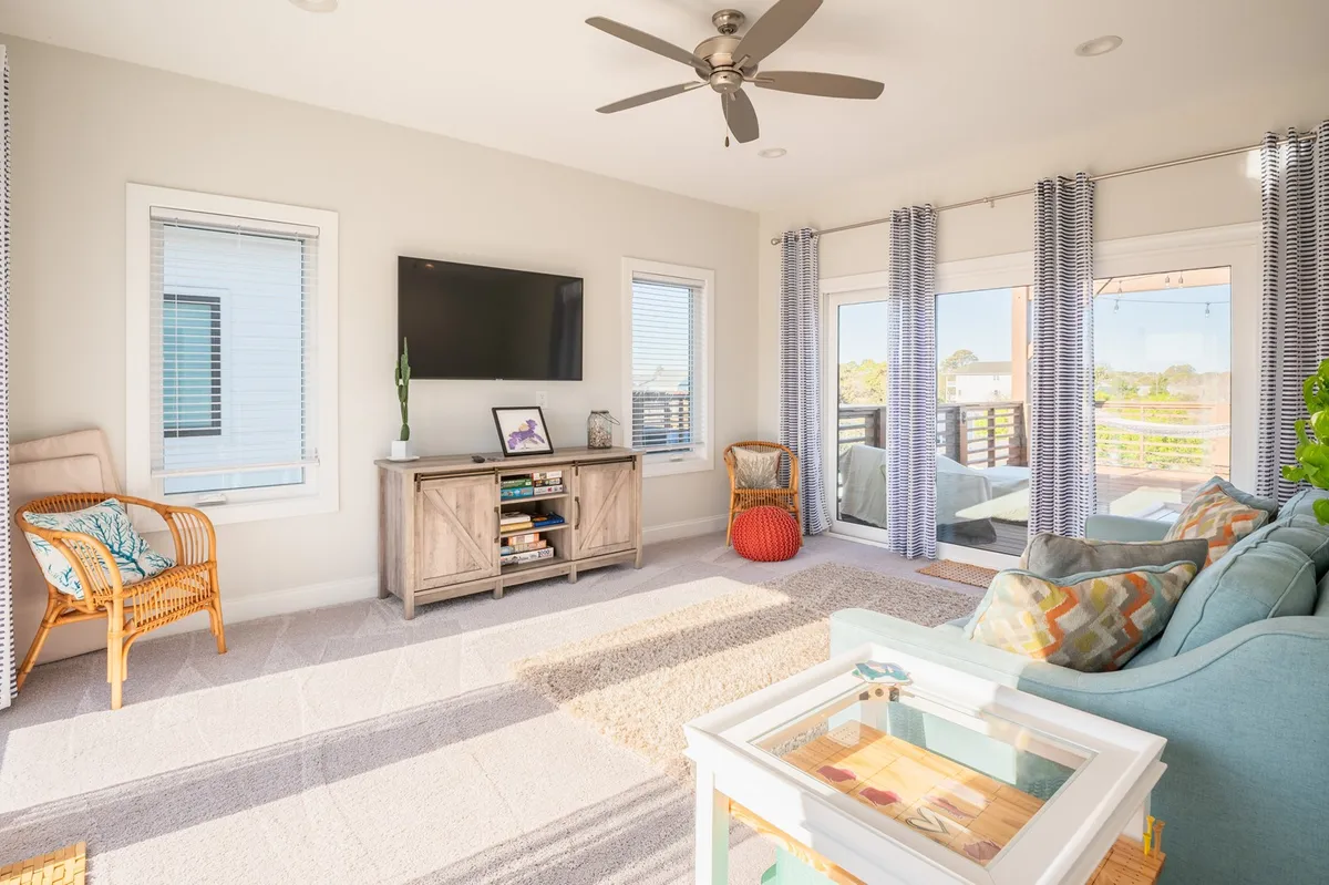 A living room featuring a plush sofa with colorful cushions, a coffee table, a large smart TV, ceiling fan, and expansive windows leading out onto the balcony. The room looks cozy for movie nights and offers a glimpse of the outdoor space.